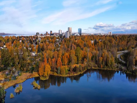 A panoramic view of Anchorage surrounded by vibrant autumn foliage, with orange and yellow trees surrounding a reflective lake in the foreground. Downtown Anchorage buildings rise in the distance under a bright blue sky with scattered clouds, and snow-capped mountains are visible on the horizon.
